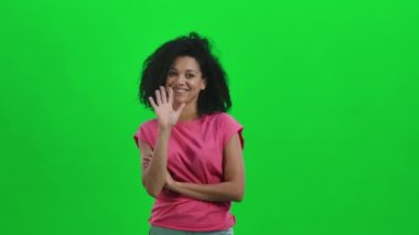 Afro-Amerikalı genç bir kadının portresi, kibarca gülümseyen, el sallayan ve el sallayan buraya gel. Kıvırcık saçlı siyah kadın yeşil ekranda poz veriyor. Kapatın. Yavaş çekim hazır 59.97fps.
