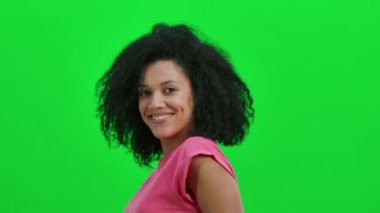 Genç Afro-Amerikan kadın portresi kameraya flört ederek gülümsüyor ve göz kırpıyor. Kıvırcık saçlı siyah kadın stüdyoda yeşil ekranda poz veriyor. Kapatın. Yavaş çekim hazır 59.97fps.