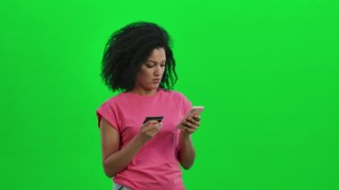 Genç Afrikalı Amerikalı kadın portresi kredi kartı ve telefonla online alışveriş yapıyor. Kıvırcık saçlı siyah kadın stüdyoda yeşil ekranda poz veriyor. Kapatın. Yavaş çekim hazır 59.97fps.