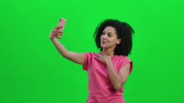 Cep telefonuyla selfie çeken genç Afrikalı Amerikalı kadın portresi. Kıvırcık saçlı siyah kadın stüdyoda yeşil ekranda poz veriyor. Kapatın. Yavaş çekim hazır 59.97fps.