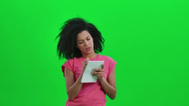 Retrato de una joven mujer afroamericana pensando, luego feliz escribiendo con un bolígrafo en un cuaderno. Mujer negra con el pelo rizado posa en la pantalla verde en el estudio. De cerca. Cámara lenta lista 59.97fps. — Vídeo de stock