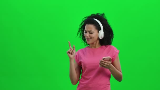 Портрет молодой женщины-афроамериканки, танцующей и наслаждающейся музыкой в больших белых наушниках с помощью смартфона. Черная женщина с вьющимися волосами позирует на зеленом экране. Закрывай. Готовность к медленному движению 59,97fps. — стоковое видео