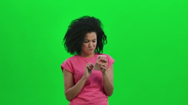 Портрет молодой афроамериканки пишет смс по телефону и радуется хорошим новостям. Черная женщина с вьющимися волосами позирует на зеленом экране в студии. Закрывай. Готовность к медленному движению 59,97fps. — стоковое видео