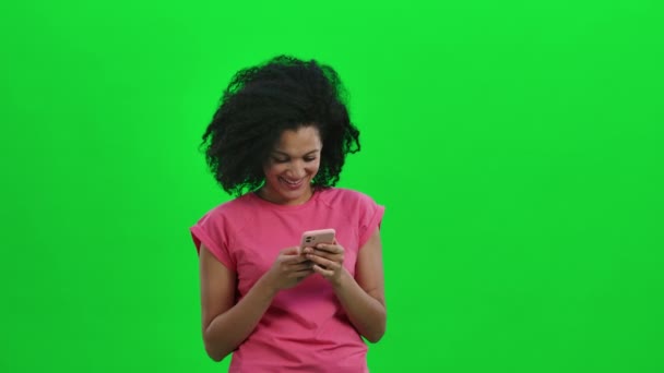 Portrét mladé Afroameričanky píše do telefonu a raduje se. Černá žena s kudrnatými vlasy pózuje na zelené obrazovce ve studiu. Zavřít. Zpomalení připraveno 59.97fps. — Stock video