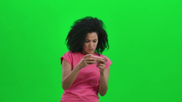 Портрет молодой афроамериканки, играющей на смартфоне и радующейся победе. Черная женщина с вьющимися волосами позирует на зеленом экране в студии. Закрывай. Готовность к медленному движению 59,97fps. — стоковое видео