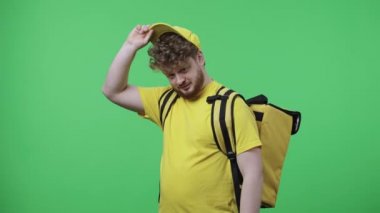 Kameraya bakıp gülümseyen genç erkeklerin portresi. Sarı kurye üniformalı, yeşil ekranda duran termo çantalı kızıl saçlı erkek. Kapatın. Yavaş çekim hazır 59.97fps.