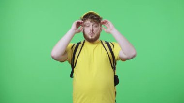 Kameraya bakan genç erkeklerin portresi şaşkınlık ve sevinçle. Sarı kurye üniformalı, yeşil ekranda duran termo çantalı kızıl saçlı erkek. Kapatın. Yavaş çekim hazır 59.97fps.