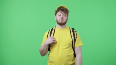 Genç yorgun adamların portresi termodinamik torba ve şapkayı çıkarıyor, mesai bitiminde. Sarı kurye üniformalı kızıl saçlı erkek stüdyoda yeşil ekranda poz veriyor. Kapatın. Yavaş çekim hazır 59.97fps.