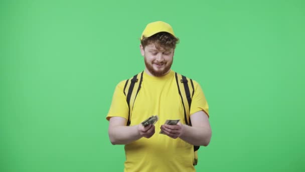 Retrato de homens jovens contando dinheiro e está muito feliz com salário ou gorjeta. Macho ruivo em uniforme de correio de entrega amarelo com saco térmico posando na tela verde. Fecha. Movimento lento pronto 59.97fps. — Vídeo de Stock