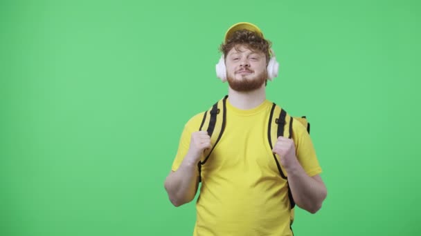 Portrét mladých mužů, kteří si užívají hudbu ve velkých bílých sluchátkách. Zrzavý muž ve žluté doručovací uniformě s termosáčkem pózující na zelené obrazovce ve studiu. Zavřít. Zpomalení připraveno 59.97fps. — Stock video