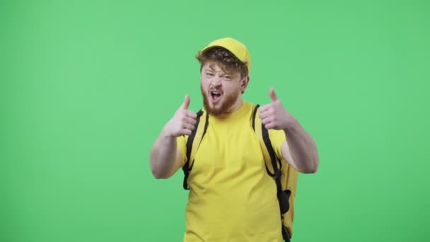 Portret młodych mężczyzn pokazujących kciuki w górę, gest jak. Rudowłosy mężczyzna w żółtym mundurze kurierskim z torbą termo pozowanie na zielonym ekranie w studio. Zamknij drzwi. Wolny ruch gotowy 59.97fps. — Wideo stockowe