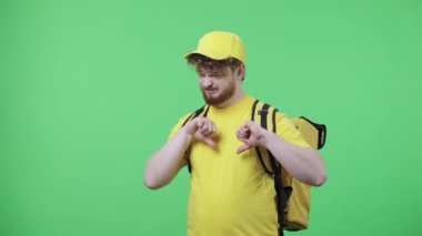 Baş parmak gösteren genç erkeklerin portresi. Sarı kurye üniformalı kızıl saçlı erkek. Stüdyoda yeşil ekranda termo çantalı poz veriyor. Kapatın. Yavaş çekim hazır 59.97fps.