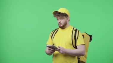 Dijital tabletten bilgi izleyen gençlerin portresi. Sarı kurye üniformalı kızıl saçlı erkek. Stüdyoda yeşil ekranda termo çantalı poz veriyor. Kapatın. Yavaş çekim hazır 59.97fps.