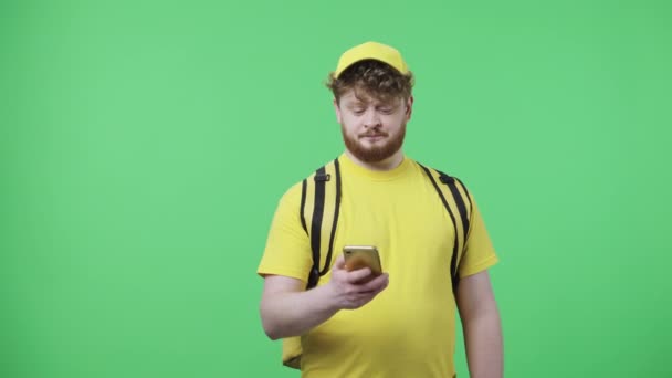 Potret pemuda yang berbicara untuk telepon, menentukan alamat pengiriman. Pria berambut merah dengan seragam kurir pengiriman kuning dengan tas termo berpose di layar hijau. Tutup. Gerakan lambat siap 59.97fps. — Stok Video