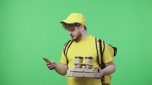 Portret mężczyzny trzymającego kawę i pizzę, podający adres dostawy za pomocą smartfona. Mężczyzna w żółtym mundurze kuriera z torbą termiczną na zielonym ekranie. Zamknij drzwi. Wolny ruch gotowy 59.97fps. — Wideo stockowe