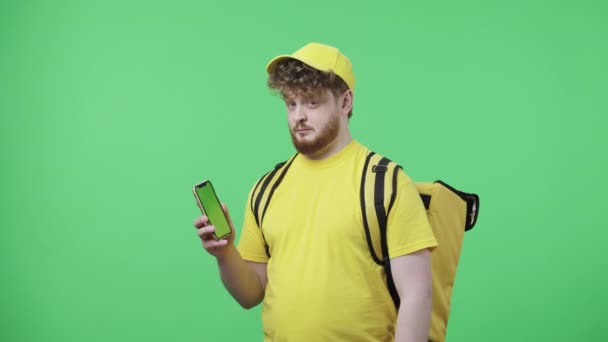 Πορτρέτο των νέων ανδρών κρατώντας τηλέφωνο με πράσινη οθόνη, δείχνει τους αντίχειρες επάνω. Redhead αρσενικό σε κίτρινη στολή κούριερ παράδοσης με θερμική τσάντα ποζάρουν στην πράσινη οθόνη. Κλείσε. Αργή κίνηση έτοιμη 59.97fps. — Αρχείο Βίντεο