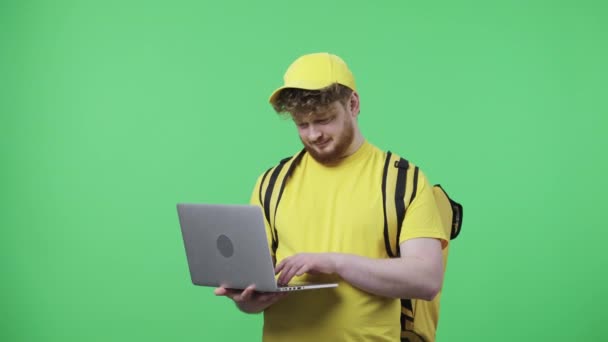 Porträt junger Männer, die auf der Laptop-Tastatur tippen. Rotschopfiges Männchen in gelber Kurieruniform mit Thermosack posiert auf grünem Bildschirm im Studio. Aus nächster Nähe. Zeitlupe bereit 59.97fps. — Stockvideo