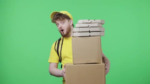 Portret młodych mężczyzn trzymających paczki i pudełka pizzy. Rudowłosy mężczyzna w żółtym mundurze kurierskim z torbą termo pozowanie na zielonym ekranie w studio. Zamknij drzwi. Wolny ruch gotowy 59.97fps. — Wideo stockowe