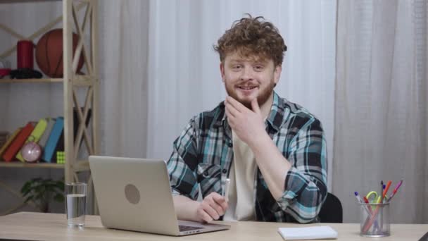 Jeune homme en chemise à carreaux regarde dans la caméra et sourit. Homme rousse avec une barbe assis dans le bureau à la maison à un bureau près d'un ordinateur portable. Ferme là. Ralenti prêt 59.97fps. — Video