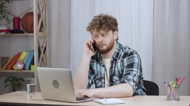 Junger Mann im karierten Hemd tippt auf einer Laptop-Tastatur und telefoniert. Der rothaarige Mann mit Bart sitzt im Home Office an einem Schreibtisch. Aus nächster Nähe. Zeitlupe bereit 59.97fps. — Stockvideo