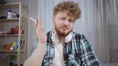 Kareli gömlekli genç adam video iletişimiyle bilgi paylaşıyor. Kızıl saçlı, sakallı bir erkek ofis masasında oturuyor. Web kamerasından görüntü. Kapatın. Yavaş çekim hazır 59.97fps.