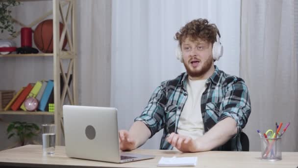 Joven con camisa a cuadros disfruta de la música con grandes auriculares blancos. Pelirrojo macho con barba sentado en la oficina de casa en un escritorio cerca de un portátil. De cerca. Cámara lenta lista 59.97fps. — Vídeos de Stock