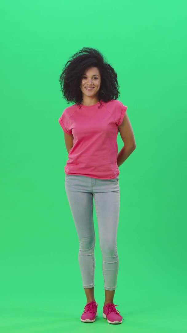 Porträtt av ung kvinna afroamerikan tittar på kameran och ler sött. Svart kvinna med lockigt hår poserar på grön skärm i studion. Slow motion klar 59,97fps. fp. Vertikal video. Full längd. — Stockvideo