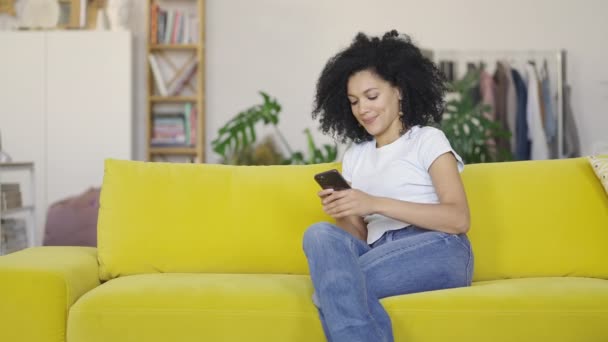 Portret młodej Afroamerykanki piszącej na telefonie. Brunetka z kręconymi włosami siedząca na żółtej kanapie w jasnym salonie. Wolny ruch gotowy 59.97fps. — Wideo stockowe