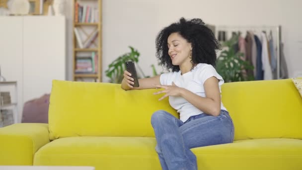 Portret młodej Afroamerykanki rozmawiającej przez telefon na smartfonie. Brunetka z kręconymi włosami siedząca na żółtej kanapie w jasnym salonie. Wolny ruch gotowy 59.97fps. — Wideo stockowe
