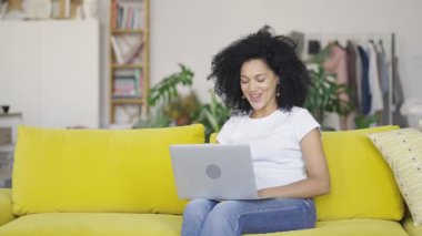 Taşınabilir bir bilgisayardan genç bir Afrikalı Amerikalı kadının portresi. Sarı koltukta oturan kıvırcık saçlı esmer, parlak bir ev odasında oturuyor. Yavaş çekim hazır 59.97fps.