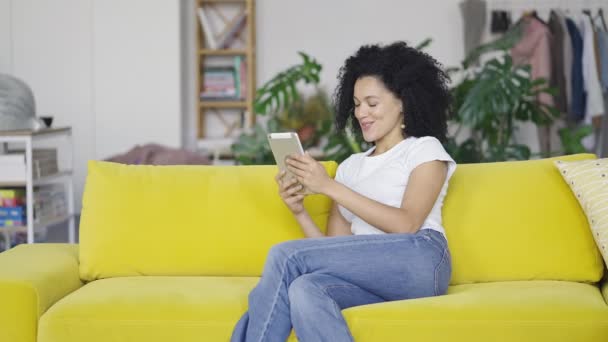 Portrét mladé Afroameričanky, která mluví na videohovoru na svém digitálním tabletu. Bruneta s kudrnatými vlasy sedí na žluté pohovce ve světlém pokoji. Zpomalení připraveno 59.97fps. — Stock video