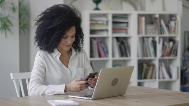 Porträt einer jungen Afroamerikanerin beim SMS-Schreiben auf einem Smartphone. Eine Brünette mit lockigem Haar in weißer Bluse sitzt an einem Tisch in einem hellen Home Office. Aus nächster Nähe. Zeitlupe bereit 59.97fps. — Stockvideo