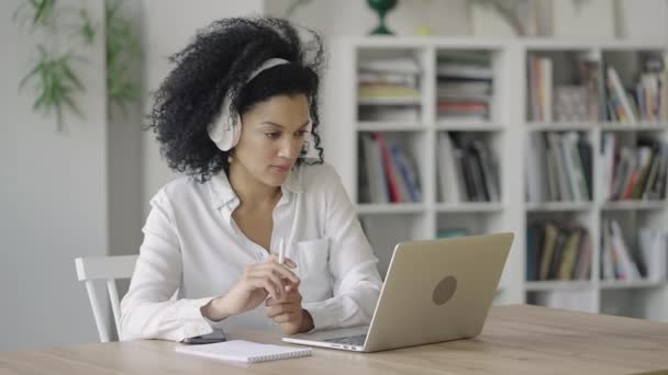 Porträt einer afroamerikanischen Frau, die in einer Videokonferenz mit Laptop und Kopfhörer Notizen auf einem Notizblock macht. Brünette sitzt am Tisch im Homeoffice. Aus nächster Nähe. Zeitlupe bereit 59.97fps. — Stockvideo