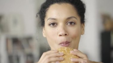 Afro-Amerikan kadın portresi. Hamburger yemek için iştahı var. Genç melez bir kadın, bulanık bir arka plan olan ışık odası karşısında fast food yiyor. Kapatın. Yavaş çekim hazır 59.97fps.
