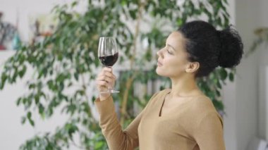 Genç Afrikalı Amerikalı kadının cam kadehten kırmızı şarap içişi. Şirin melez kadın ışık odasının arka planında içkinin tadını çıkarıyor. Kapatın. Yavaş çekim hazır 59.97fps.