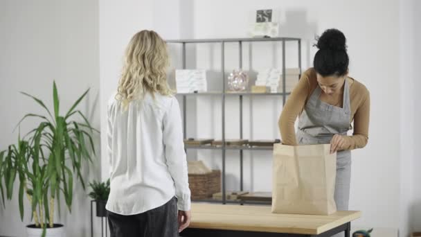 Jeune fille ramasse un sac en papier avec sa commande en ligne dans un magasin. Une femme afro-américaine mignonne emballe des marchandises pour un client dans le contexte de la pièce lumineuse. Ralenti prêt 59.97fps. — Video