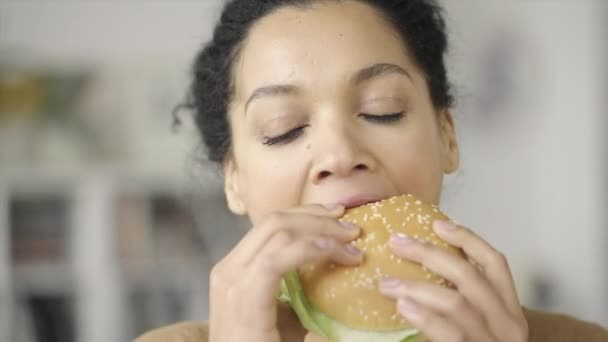 Potret wanita Afrika-Amerika dengan nafsu makan untuk menggigit hamburger. Perempuan ras campuran muda makan makanan cepat saji dengan latar belakang kabur dari ruang cahaya. Tutup. Gerakan lambat siap 59.97fps. — Stok Video
