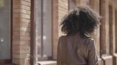 Genç Afro-Amerikan kadın sokakta yürürken, arkasını dönüyor ve gülümsüyor. Kahverengi deri ceketli esmer, bulanık tuğla binaya karşı duruyor. Kapatın. Yavaş çekim hazır 59.97fps.