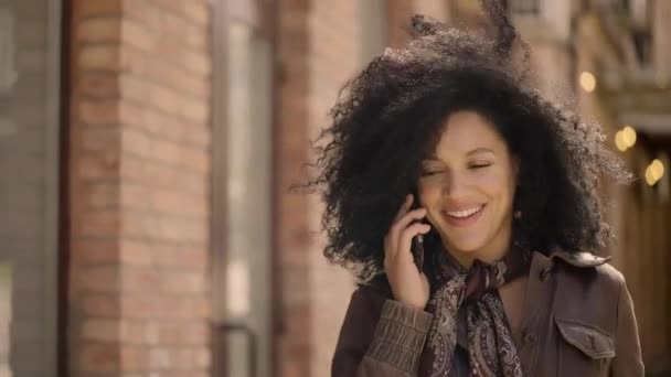 一个年轻的非洲裔美国女人用智能手机说话的画像。穿着皮夹克卷曲头发的布鲁内特在模糊的砖楼背景下走在大街上。靠近点慢动作准备59.97fps — 图库视频影像