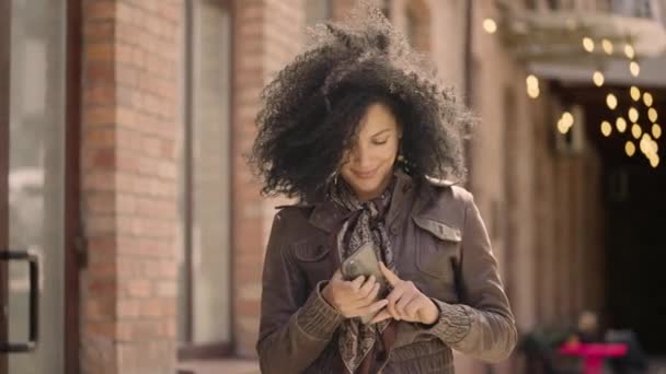 年轻的非洲裔美国女人用智能手机发短信的肖像。穿着皮夹克的布鲁内特在模糊的砖楼背景下沿街走着。靠近点慢动作准备59.97fps. — 图库视频影像