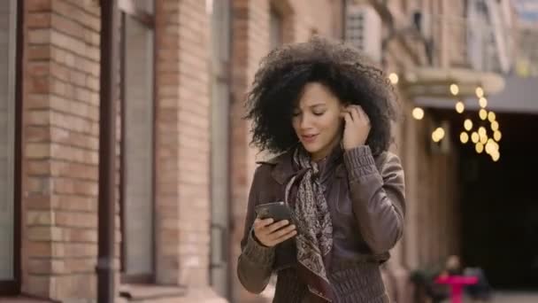 非洲裔美国女青年用智能手机用无线耳机跳舞和欣赏音乐的肖像。穿着皮夹克的布鲁内特走在街上.靠近点慢动作准备59.97fps. — 图库视频影像
