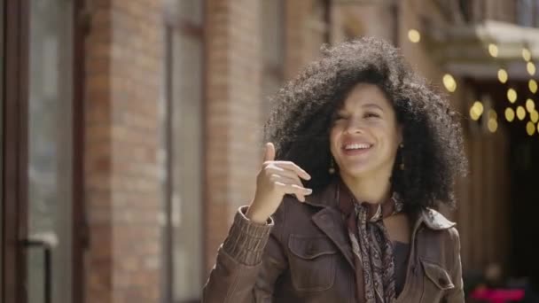 Portret młodej Afroamerykanki machającej ręką, pokazującej gest. Brunetka w brązowej skórzanej kurtce pozująca na ulicy przed zamazanym murowanym budynkiem. Zamknij drzwi. Wolny ruch gotowy 59.97fps. — Wideo stockowe
