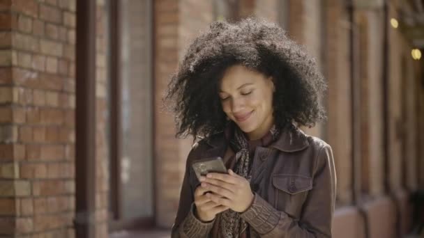 年轻的非洲裔美国女人用智能手机发短信的肖像。穿着皮夹克的布鲁内特在模糊的砖楼背景下沿街走着。靠近点慢动作准备59.97fps. — 图库视频影像