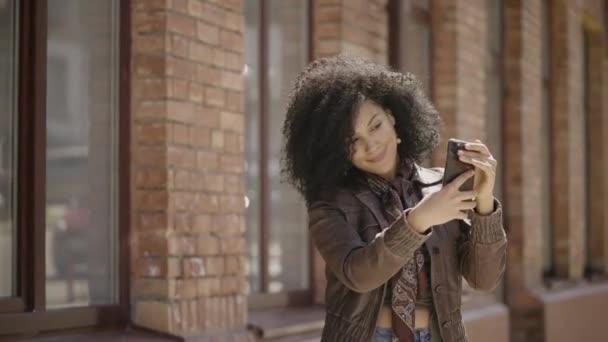 Портрет стильной молодой афроамериканской женщины, делающей селфи на мобильном телефоне, затем смотрящей фотографии. Брюнетка в коричневой кожаной куртке позирует на улице. Закрывай. Готовность к медленному движению 59,97fps. — стоковое видео