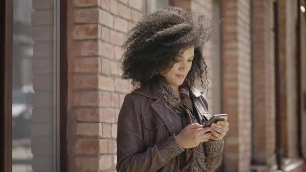 Портрет стильной молодой афроамериканки, смотрящей информацию по телефону. Брюнетка в коричневой кожаной куртке позирует на улице против размытого кирпичного здания. Закрывай. Готовность к медленному движению 59,97fps. — стоковое видео