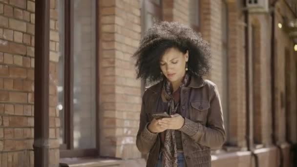 Портрет стильной молодой афроамериканки, смотрящей информацию по телефону. Брюнетка в коричневой кожаной куртке идет по улице против размытого кирпичного здания. Закрывай. Готовность к медленному движению 59,97fps. — стоковое видео