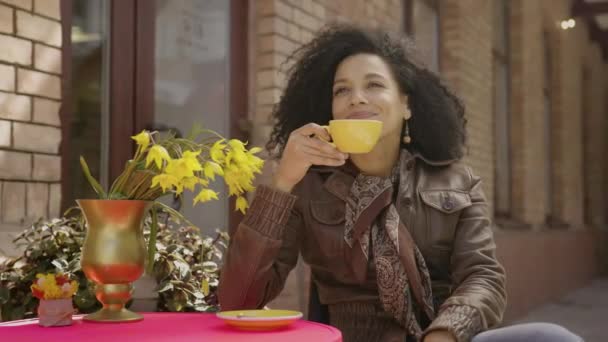 Porträt einer jungen Afroamerikanerin, die das Aroma und den Geschmack von heißem Kaffee genießt. Brünette in brauner Lederjacke sitzt am Tisch in einem gemütlichen Café auf der Straße. Aus nächster Nähe. Zeitlupe bereit 59.97fps. — Stockvideo