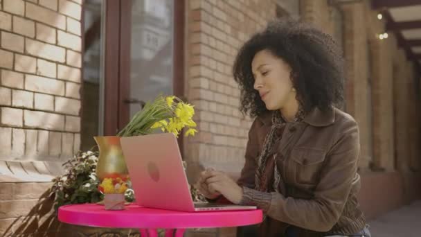Porträt einer eleganten jungen Afroamerikanerin, die per Videotelefonie mit einem Laptop spricht. Brünette in brauner Lederjacke sitzt an einem Tisch in einem gemütlichen Café auf der Straße. Aus nächster Nähe. Zeitlupe bereit 59.97fps. — Stockvideo