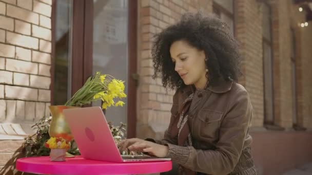 아프리카 계 미국인젊은 여성 이 노트북 키보드로 타이핑을 하고 커피를 마시는 모습. 갈색 가죽 재킷을 입은 브루넷은 거리의 아늑 한 카페 탁자에 앉아 있었다. 닫아. 느린 동작 준비 59.97fps. — 비디오