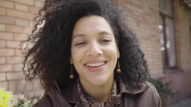 Retrato da jovem afro-americana falando em videochamada. Morena em jaqueta de couro marrom posando na rua contra edifício borrado. Vista da câmera web. Fecha. Movimento lento pronto 59.97fps. — Vídeo de Stock